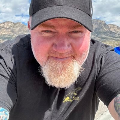 A close-up selfie of author John Tackett wearing a black baseball cap and black t-shirt, smiling outdoors with a mountainous landscape in the background.